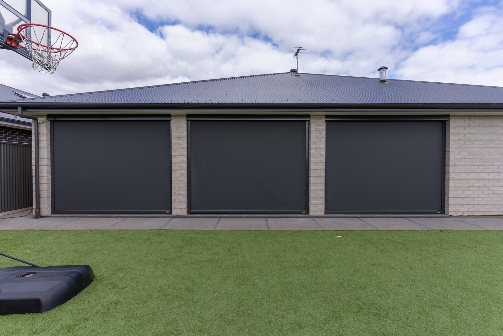 Three dark outdoor blinds fully lowered across the alfresco of a brick home, with lawn in front and open sky above. Three dark outdoor blinds fully lowered across the alfresco of a brick home, with lawn in front and open sky above.