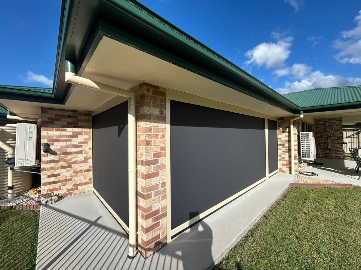 Charcoal track-guided outdoor blinds closed between brick piers, the fabric edges held in cream side channels